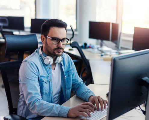 Side view of IT pprogrammer sitting at computer with headphones around his neck